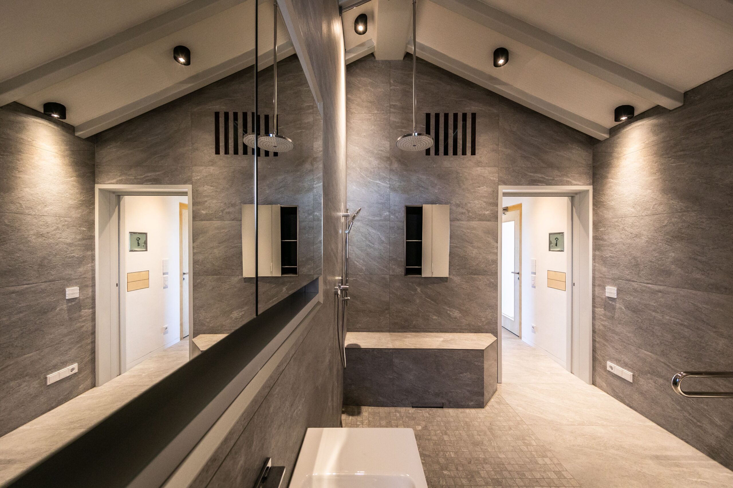 Modernes Badezimmer mit grauen Fliesen, großer Spiegelwand, Dusche und zwei offenen Türen zu angrenzenden Räumen.