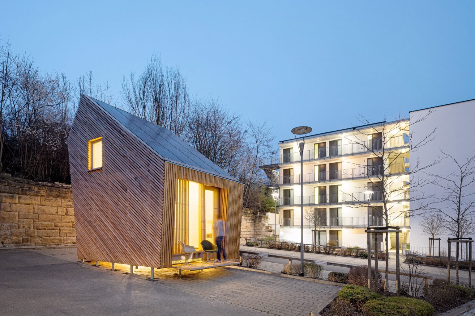 Tiny House Außenansicht bei Dämmerung neben weißen Gebäude.