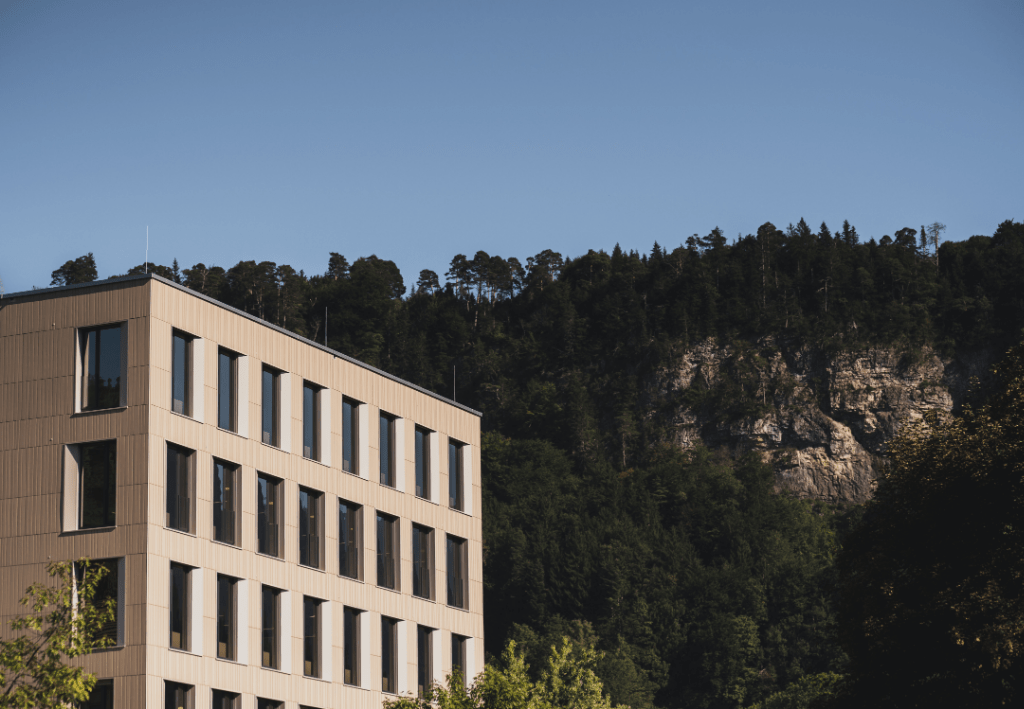 Helle Keramikfassade mit Wald im Hintergrund.