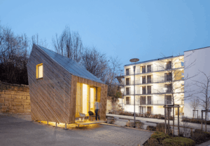 Tiny House Außenansicht bei Dämmerung neben weißen Gebäude.