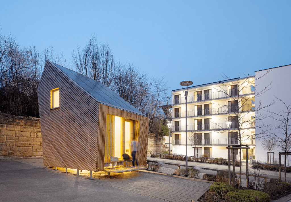 Tiny House Außenansicht bei Dämmerung neben weißen Gebäude.