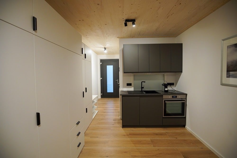 Kleine moderne Küchenzeile mit grauen Schränken, Backofen und Spüle in einem hellen Raum mit Holzboden und weißen Wänden.