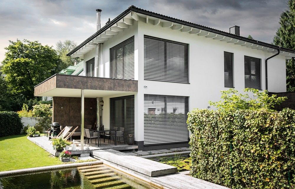 Modernes zweistöckiges weißes Haus mit großen Fenstern und Außenjalousien, umgeben von Garten und Teich.