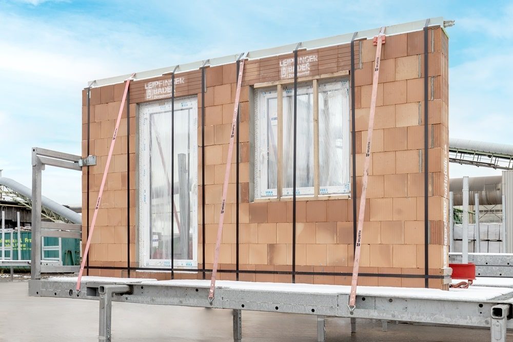 Fertige Ziegelwand mit zwei weißen Fensterzargen, befestigt mit Gurten auf einer Metallplattform.