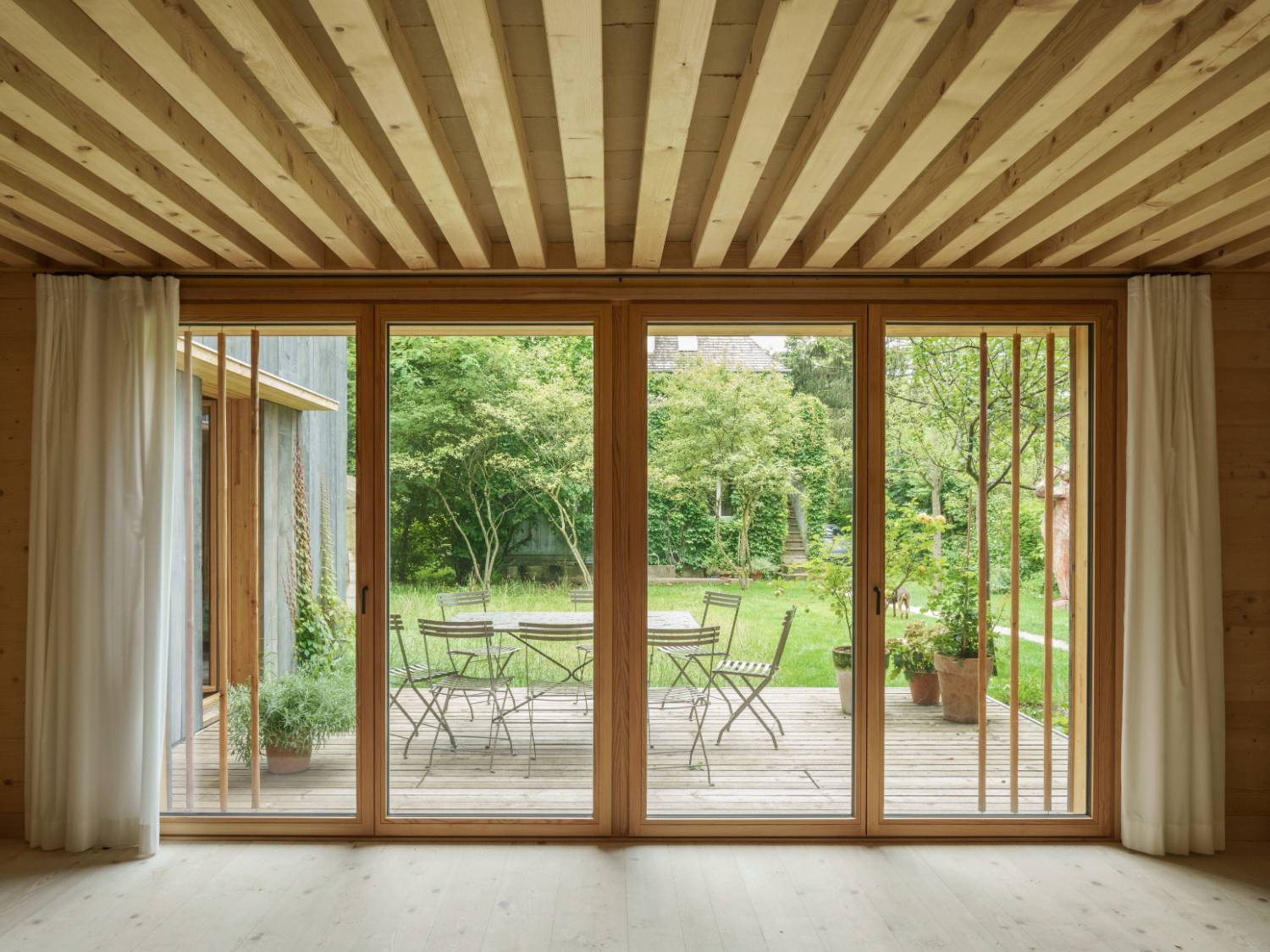 Blick durch große Holzrahmen-Schiebetüren auf Terrasse mit Metalltisch und Stühlen, grüner Garten im Hintergrund.