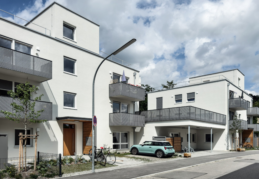 Hybrid genutzter Neubau in Landshut mit Leipfinger-Bader Systemlösungen. Weißer Fassade, mehreren Balkonen, geparktem Auto und Fahrrädern vor dem Gebäude.