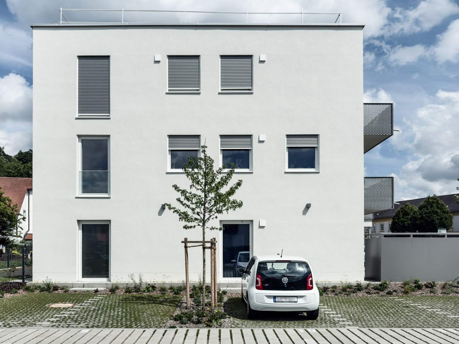 Hybrid genutzter Neubau in Landshut mit weißer Fassade, mehreren Fenstern, kleinem Baum davor und einem geparkten weißen Auto auf Parkplatz.