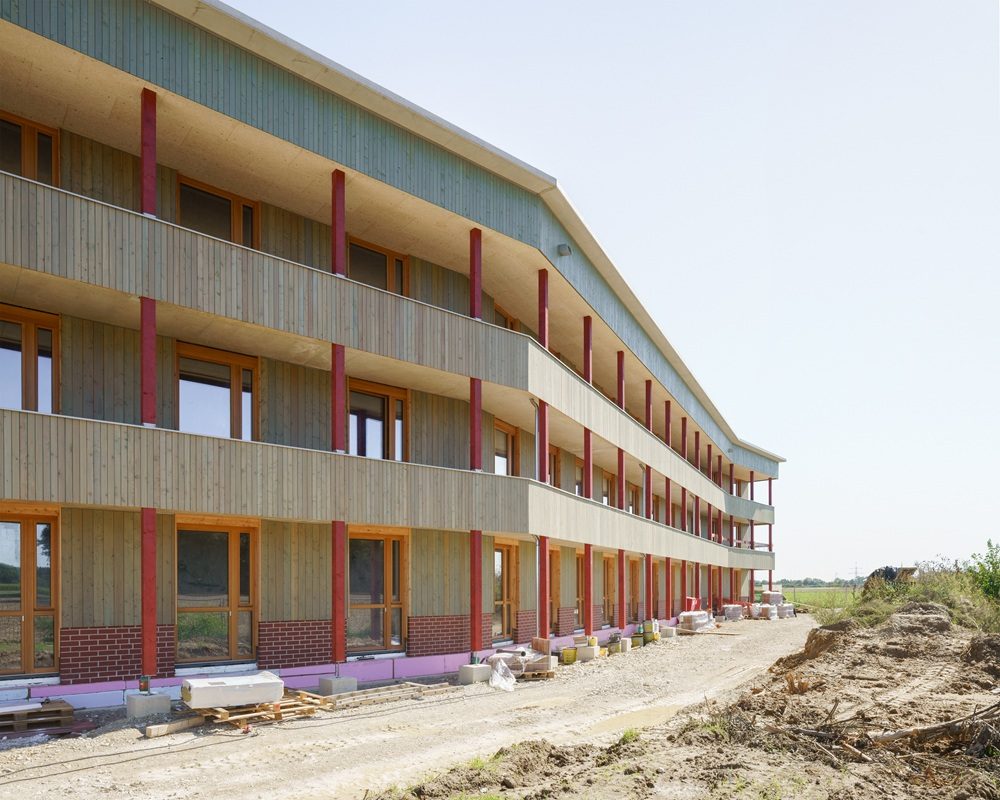 Haus fast ohne Heizung in Ingolstadt: Mehrstöckiges Wohngebäude mit Holzfassade und roten Stützen, Baustelle mit Erde und Baumaterialien im Vordergrund, klarer Himmel.