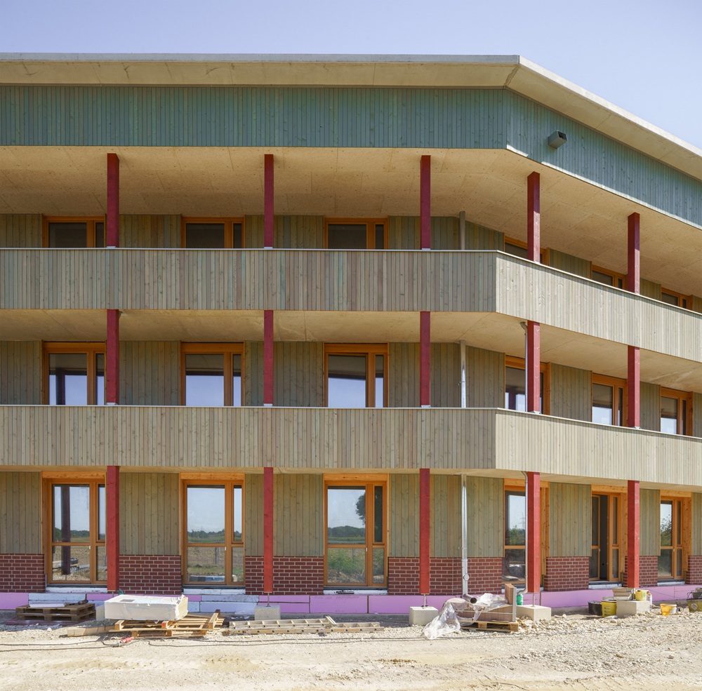 Haus fast ohne Heizung in Ingolstadt: Mehrstöckiges Holzgebäude mit großen Fenstern und Balkonen, Baustelle mit Baumaterialien im Vordergrund.
