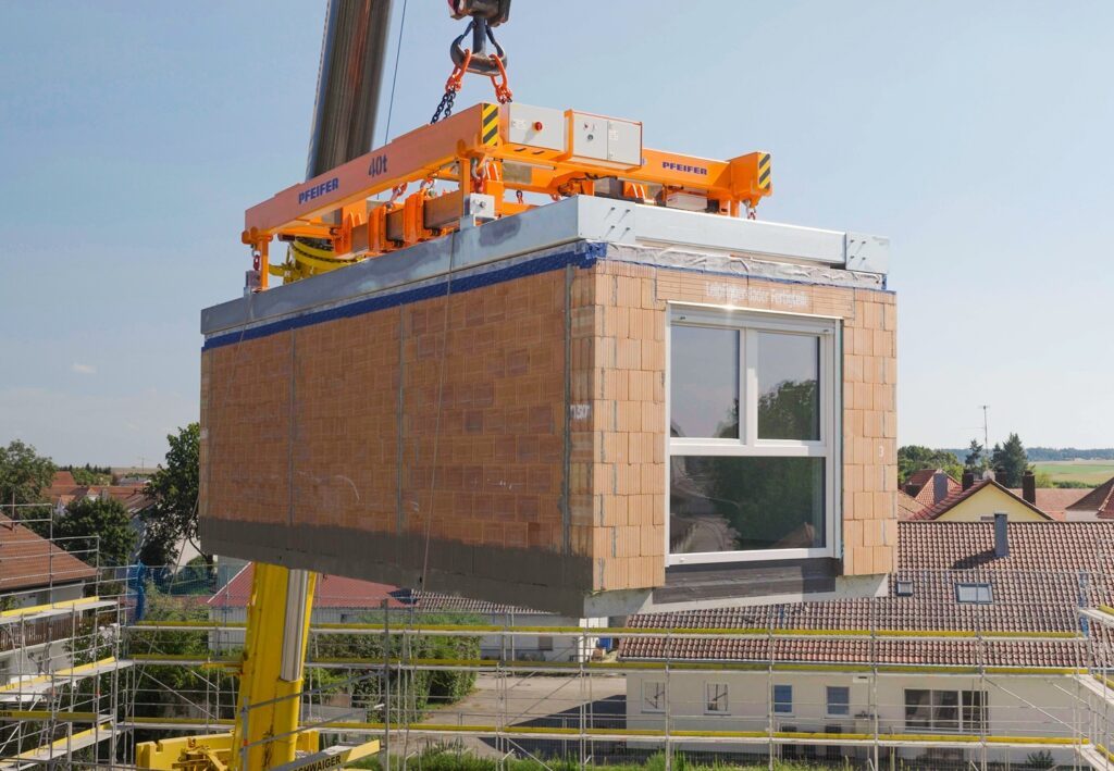 Vorgefertigtes Ziegelmodul mit Fenster, das an einem Kranhaken hängt, im Hintergrund Häuser und blauer Himmel.
