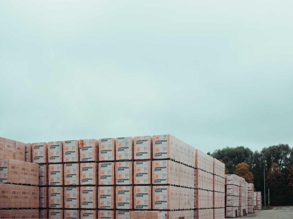 Mehrere Paletten mit verpackten Ziegelsteinen der Marke Leipfinger-Bader, gestapelt im Freien vor Bäumen.