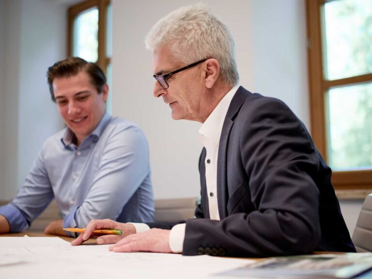 Zwei Männer sitzen an einem Tisch, einer trägt ein schwarzes Sakko und ein weißes Hemd, der andere ein hellblaues Hemd.