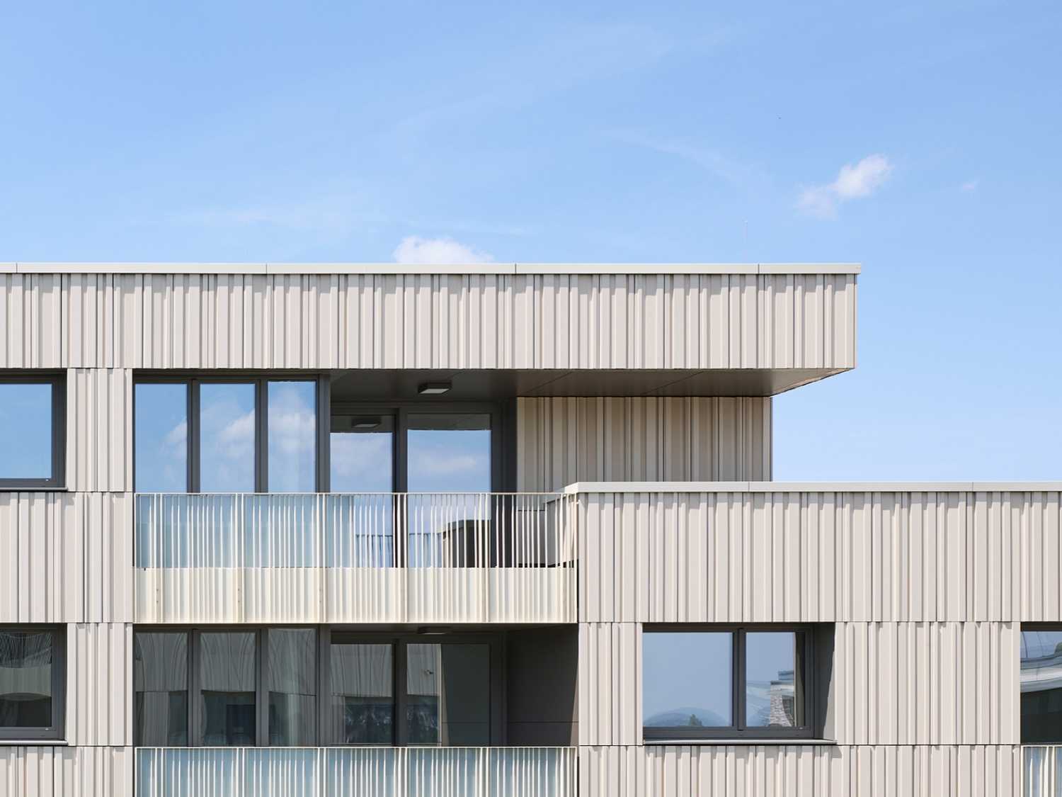 Moderner Gebäudefassade mit vertikal strukturierten, hellen Keramikplatten und großen Fenstern, darunter ein Balkon mit Glasgeländer.