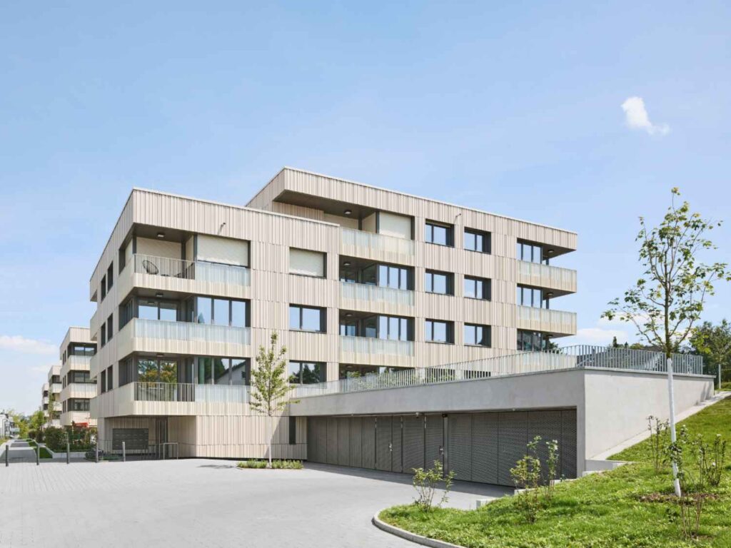 Modernes Wohngebäude mit Ton-in-Ton Keramikfassade in Pforzheim, rechteckige Fenster, flache Dächer, davor Garagen mit Metalltoren und Grünflächen mit jungen Bäumen.