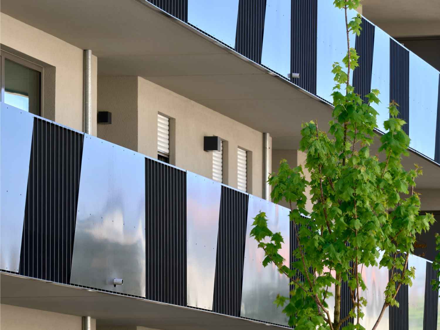 Mehrstöckiges Wohngebäude mit Balkonen, die abwechselnd mit glatten und geriffelten Metallplatten verkleidet sind, und einem Baum im Vordergrund.
