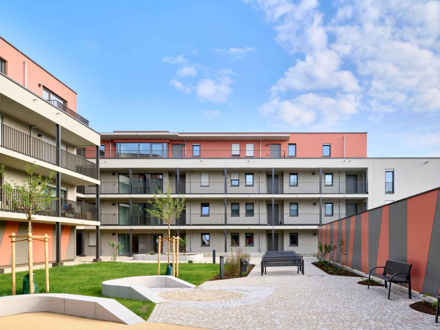Moderner Wohnkomplex mit drei Etagen, Balkonen, gepflastertem Innenhof, Sitzbänken und jungen Bäumen.