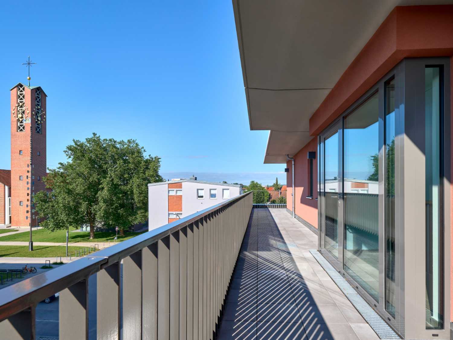 Balkon mit Metallgeländer an einem modernen Gebäude, im Hintergrund ein Kirchturm und Bäume unter blauem Himmel.