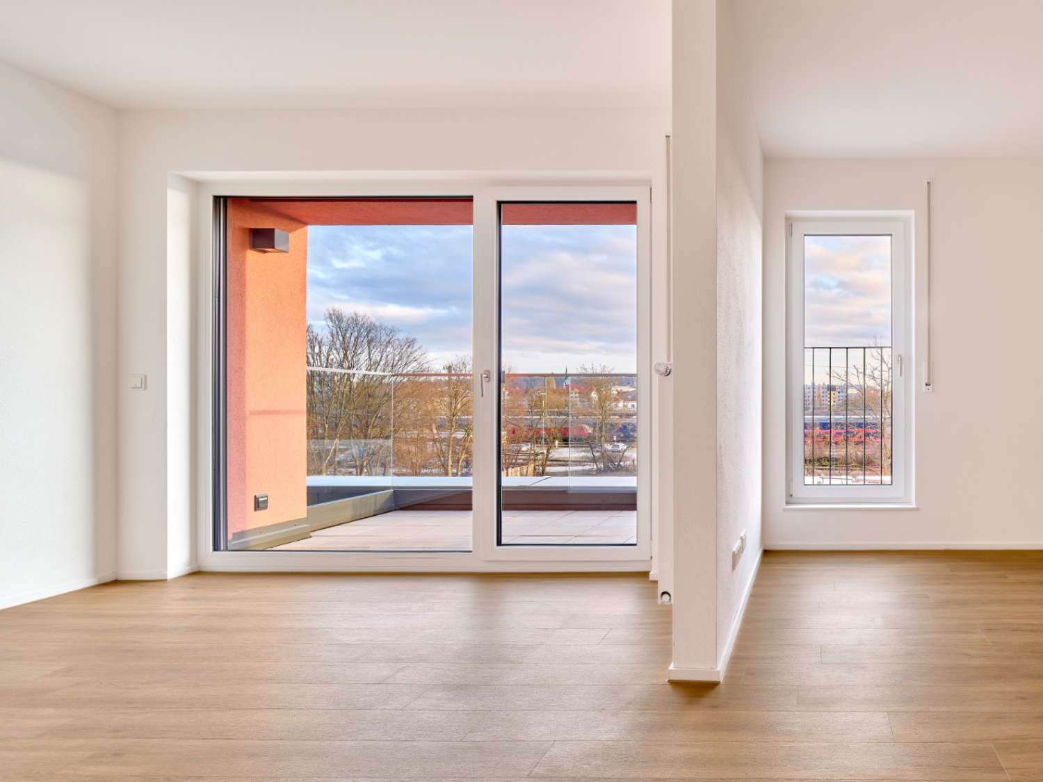 Leerer Raum mit hellen Wänden, großem bodentiefem Fenster und kleinem Fenster mit Blick auf Bäume und Gebäude.