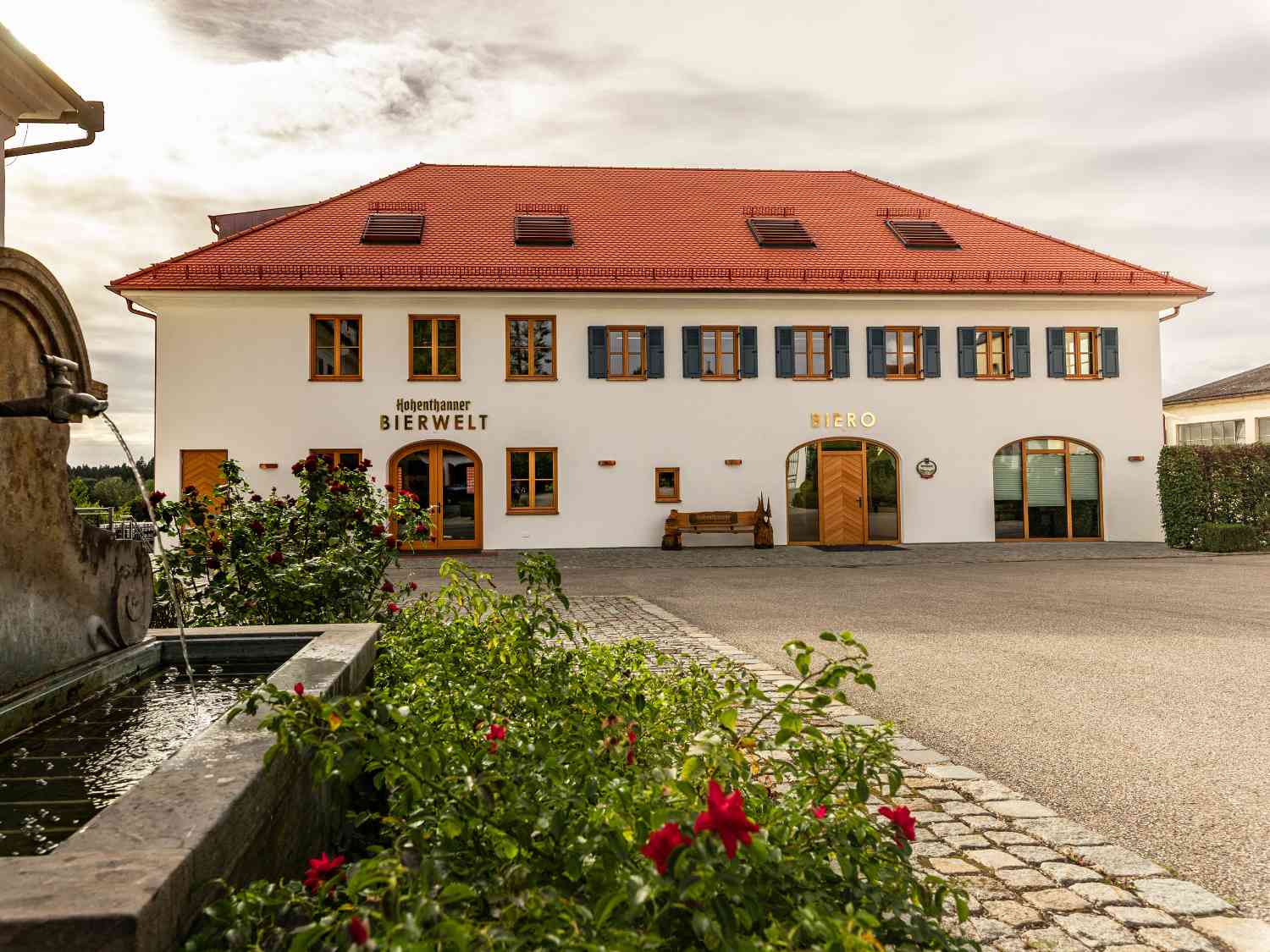 Zweistöckiges weißes Gebäude mit rotem Ziegeldach, mehreren Fenstern mit blauen Fensterläden, den Schriftzügen 'Bierwelt' und 'Biero' an der Fassade sowie einem Brunnen und Blumen im Vordergrund.