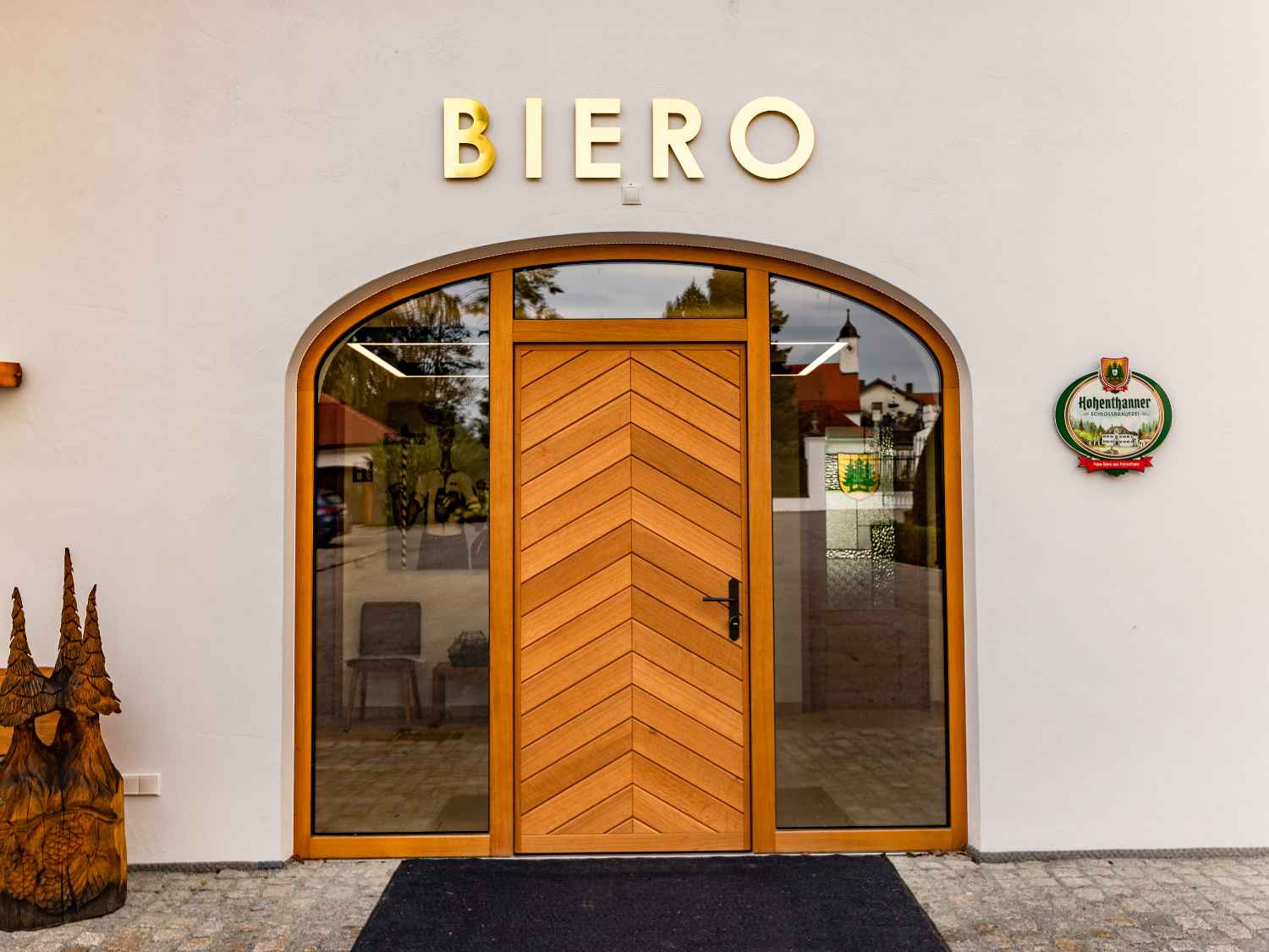 Eingang mit hölzerner Tür und zwei großen Fenstern, darüber die goldfarbene Aufschrift 'BIERO', rechts ein rundes Schild mit dem Schriftzug 'Hohenthanner'.