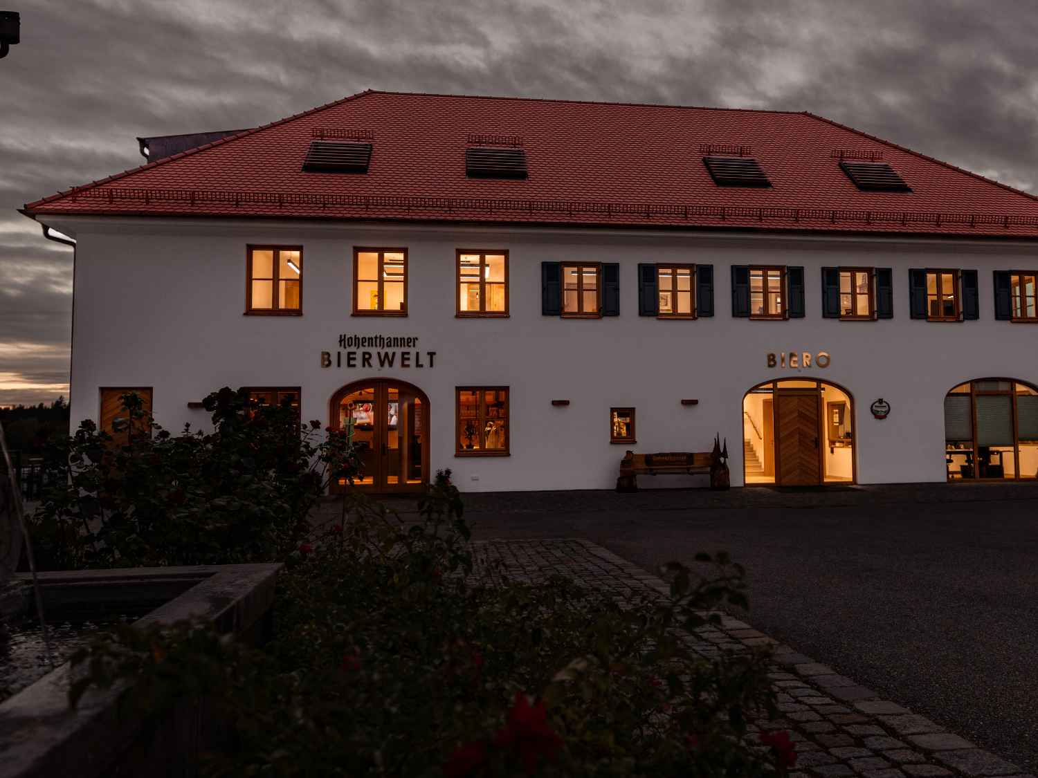 Zweistöckiges Gebäude mit rotem Satteldach, beleuchteten Fenstern und den Schriftzügen 'Hohenthanner BIERWELT' und 'BIERO' an der Fassade bei Dämmerung.