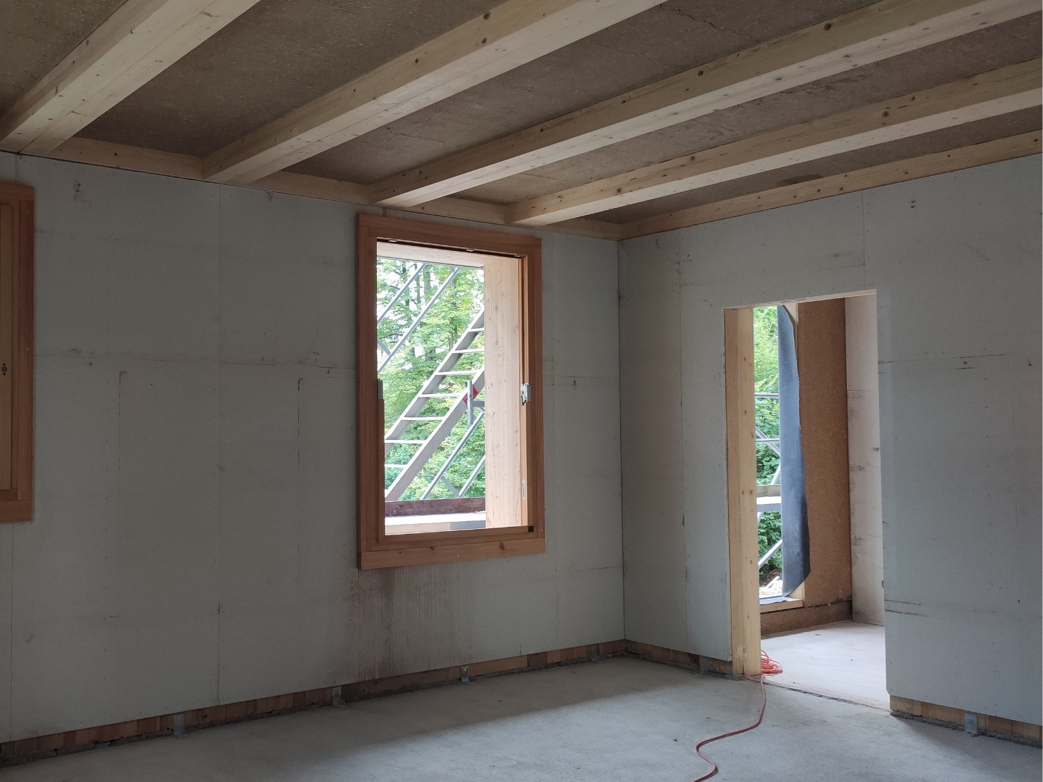 Innenraum eines Rohbaus mit sichtbaren Holzträgern der Leipfinger-Bader Holz-Lehm Massivdecke, zwei Fenster mit Holzrahmen.