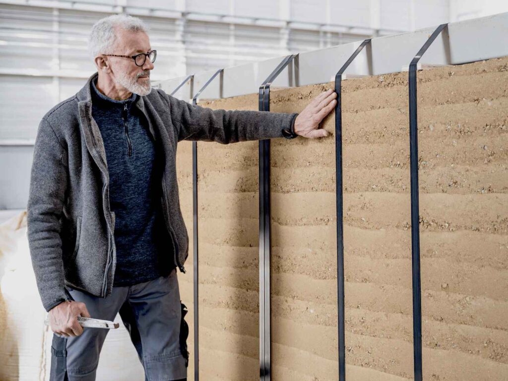 Ein Mann steht neben einer Stampflehmfertigteil von Leipfinger-Bader und berührt diese mit der Hand, er hält ein Werkzeug in der anderen Hand.