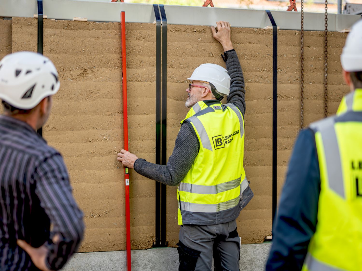 Drei Personen mit Schutzhelmen und gelben Warnwesten mit Aufdruck 'Leipfinger Bader' stehen vor einer Stampflehmwand, eine Person misst die Wand mit einer langen roten Stange.