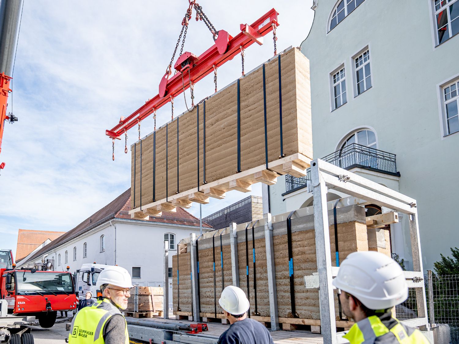 Mehrere Bauarbeiter mit Schutzhelmen heben mit einem Kran große, rechteckige Stampflehmwand-Elemente auf einer Baustelle neben einem historischen Gebäude.