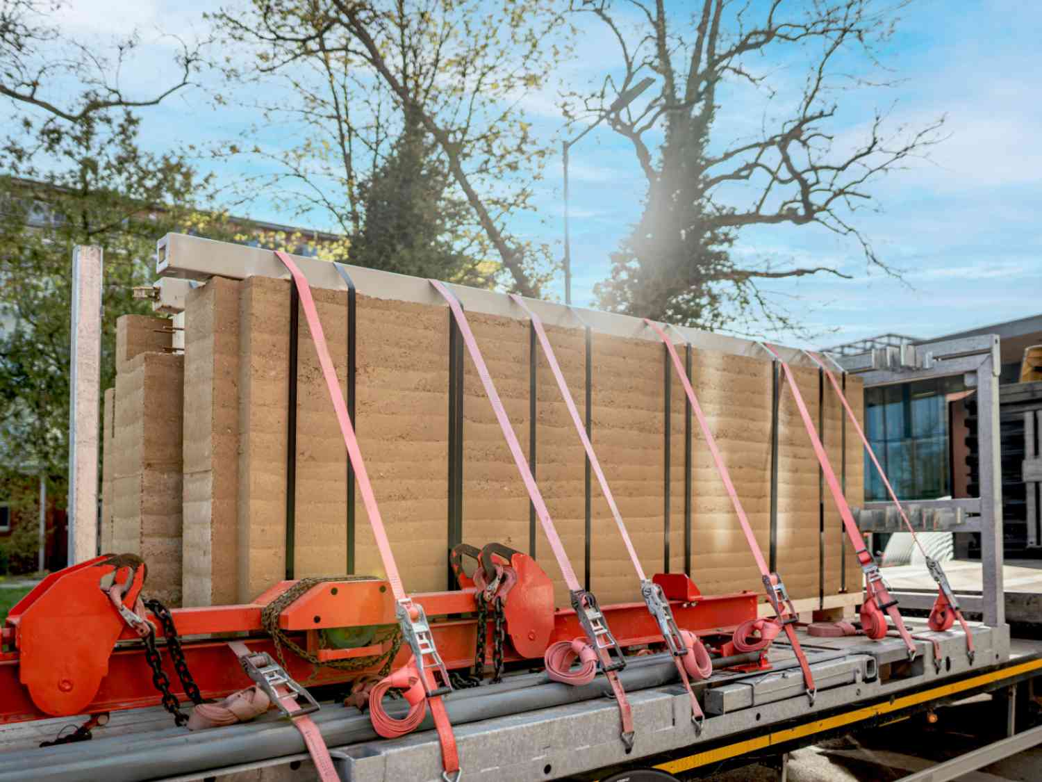 Stampflehmwand auf einem Lkw mit mehreren roten Spanngurten gesichert, im Hintergrund Bäume und ein Gebäude.