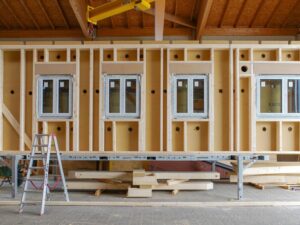 Vorgefertigte Holzwand mit vier Fensterausschnitten und eingebauten Fenstern, davor eine Leiter und ein Arbeitstisch mit Holzlatten.